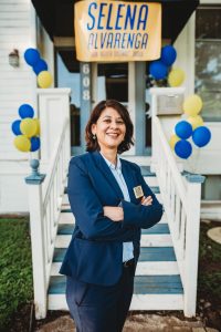 Selena Alvarenga stands facing the camera with arms crossed in front of a campaign sign.